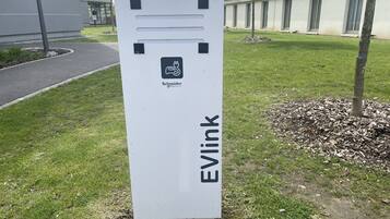 A white EVlink electric vehicle charging station with a logo, located outdoors in a grassy area with a building in the background.
