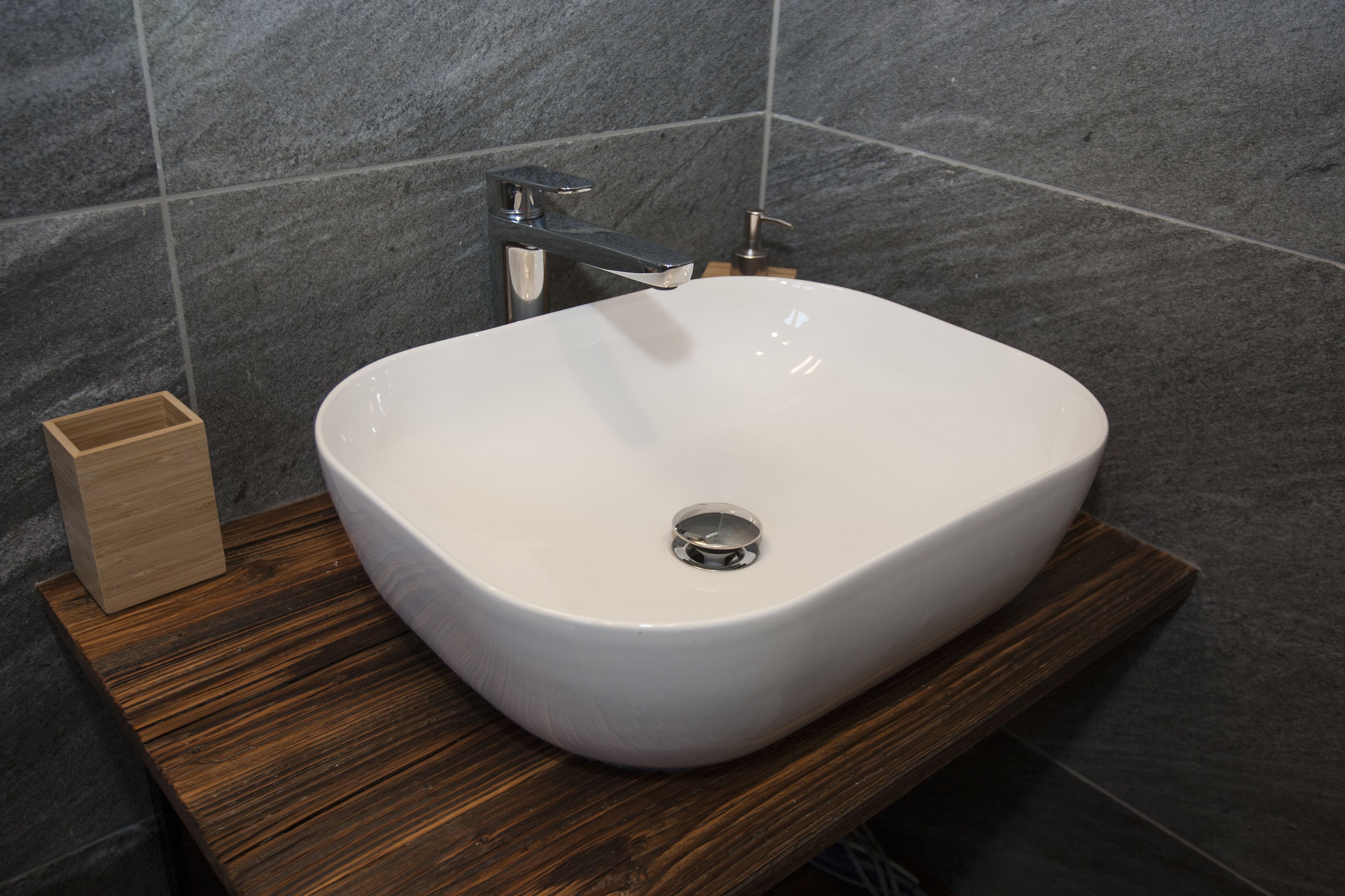 Un bagno con lavandino bianco, mobile contenitore in legno e pareti rivestite di piastrelle scure.
