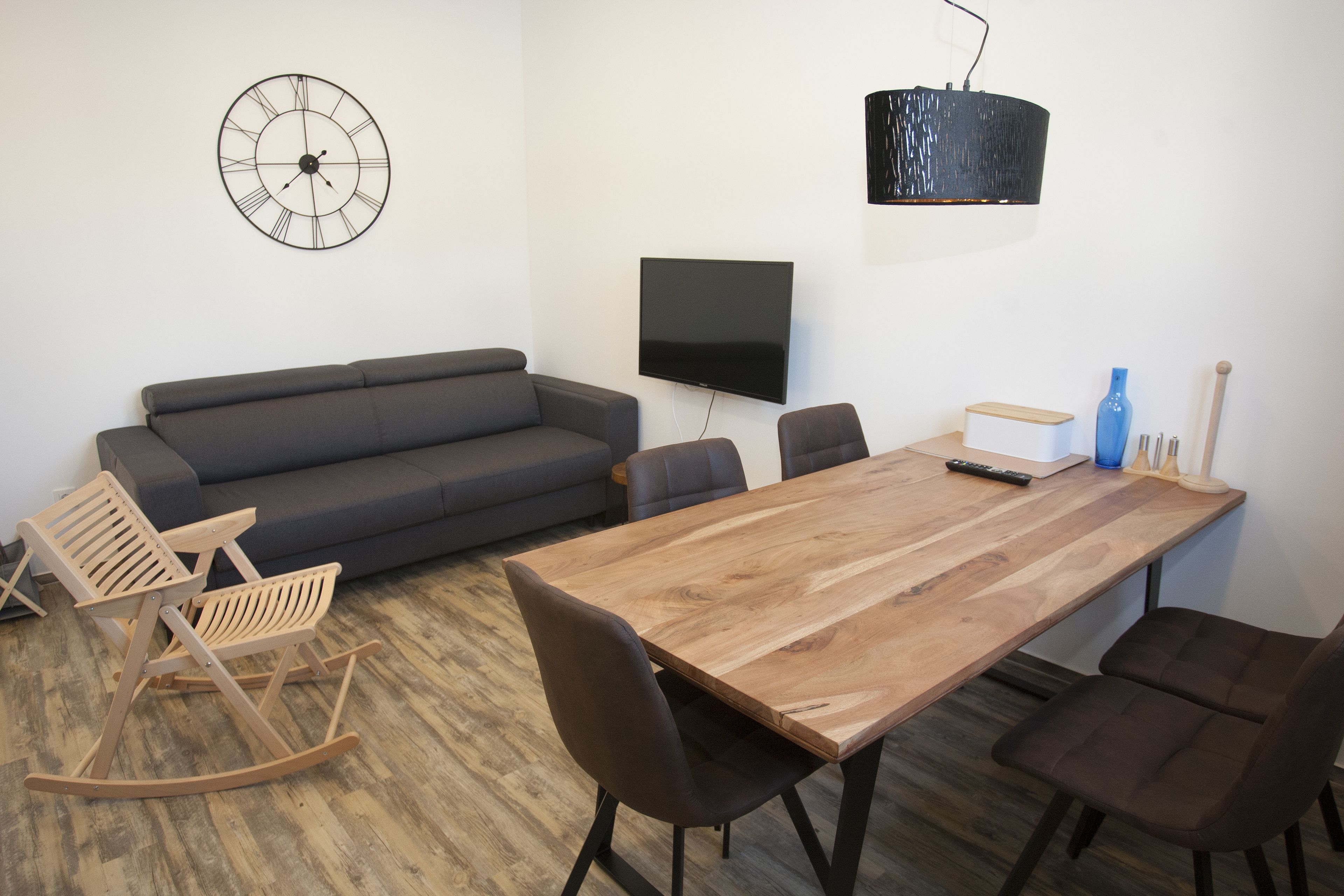 Una sala moderna con una mesa de comedor de madera, un sofá gris oscuro y una mecedora de madera. Un televisor de pantalla plana está montado en la pared, y una lámpara colgante negra ilumina la mesa.