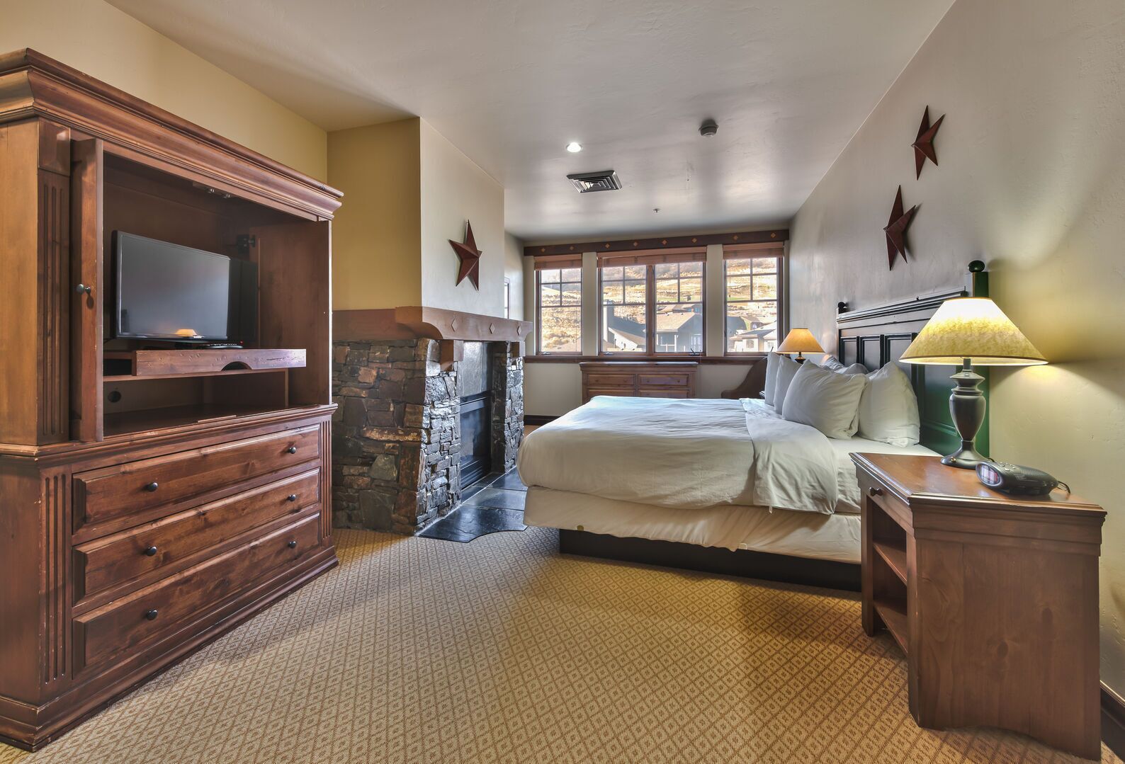 Une chambre avec une cheminée en pierre, une grande commode en bois, un lit avec du linge de maison blanc, une table de chevet avec une lampe, et une vue sur l’extérieur par une fenêtre.