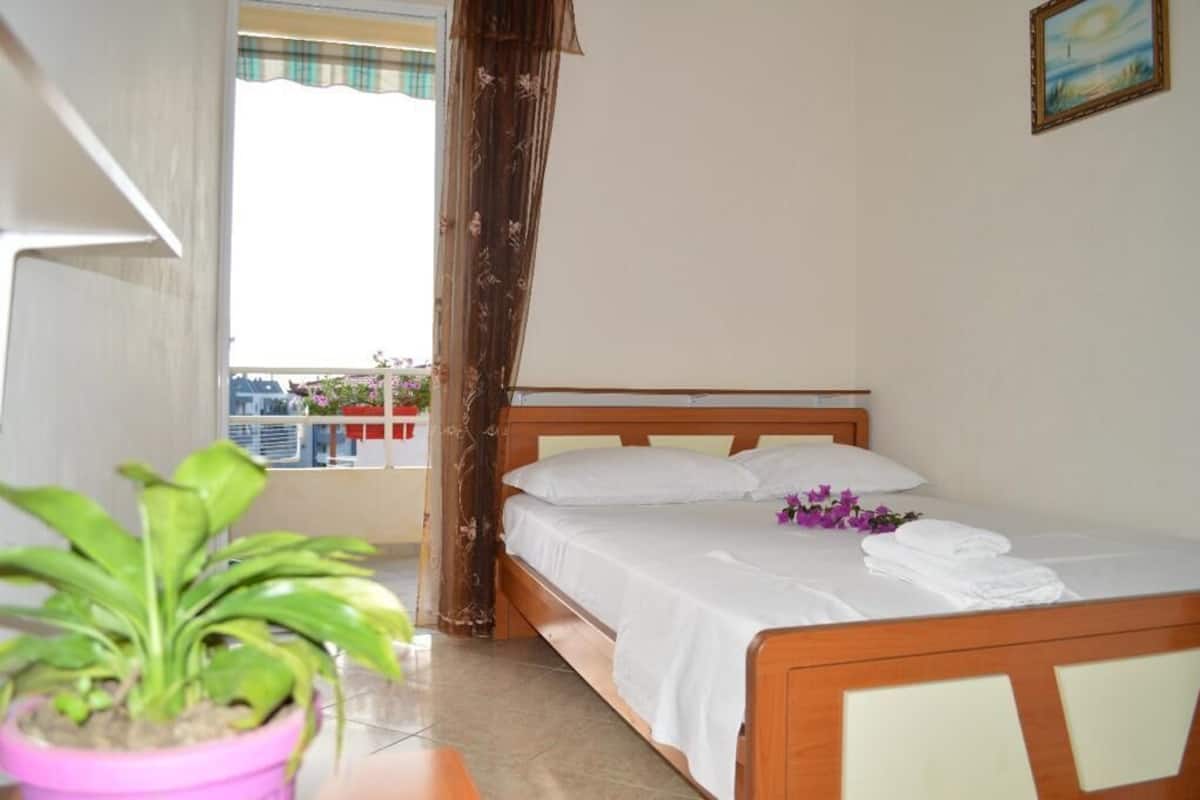 Habitación de hotel con cama de madera, ropa de cama blanca, una planta en maceta y vistas de la ciudad a través de una ventana con cortina.