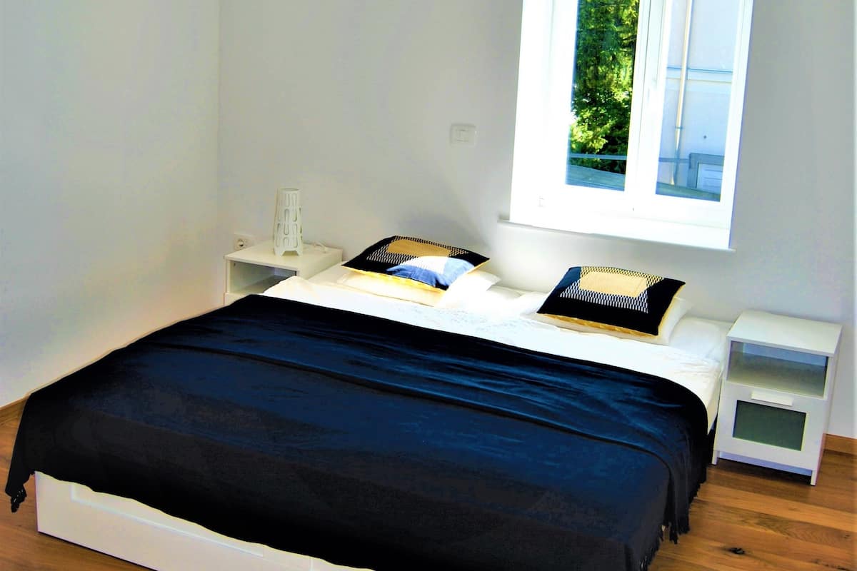 A bedroom with a large window, a bed with a black bedspread, white nightstands, and a white wall.