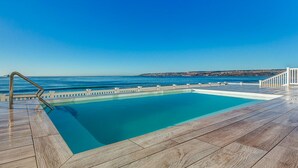 En swimmingpool med ett trädäck och ett metallräcke, med utsikt över en vattenyta och en avlägsen kuststräcka.