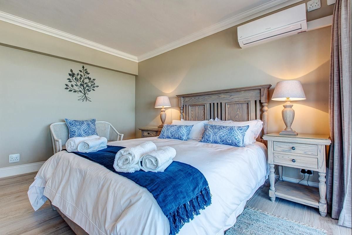 A neatly made bed with white and blue bedding, two bedside tables with lamps, a wall-mounted air conditioner, and a decorative wall piece.