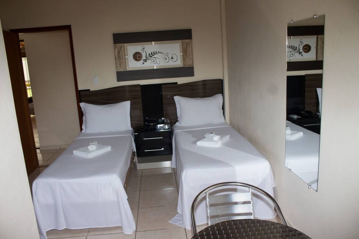 A hotel room with two single beds, a bedside table with a lamp, a small table with a chair, and a large mirror on the wall.