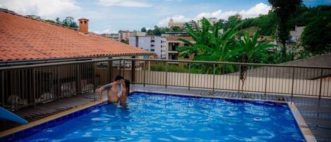 Una piscina con borde de baldosas, rodeada por una cerca metálica y con vistas a edificios y follaje al fondo.