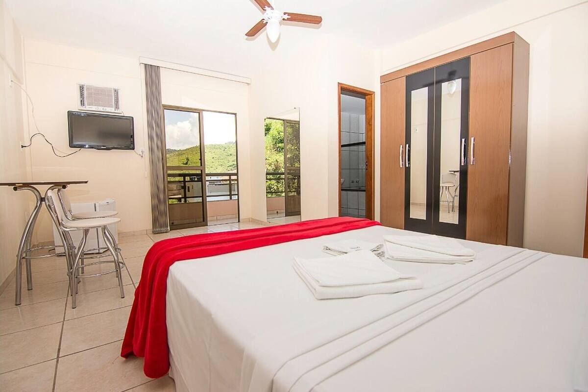 A hotel room with a bed, a desk with a chair, a TV, a wardrobe, and a balcony with a view of greenery.