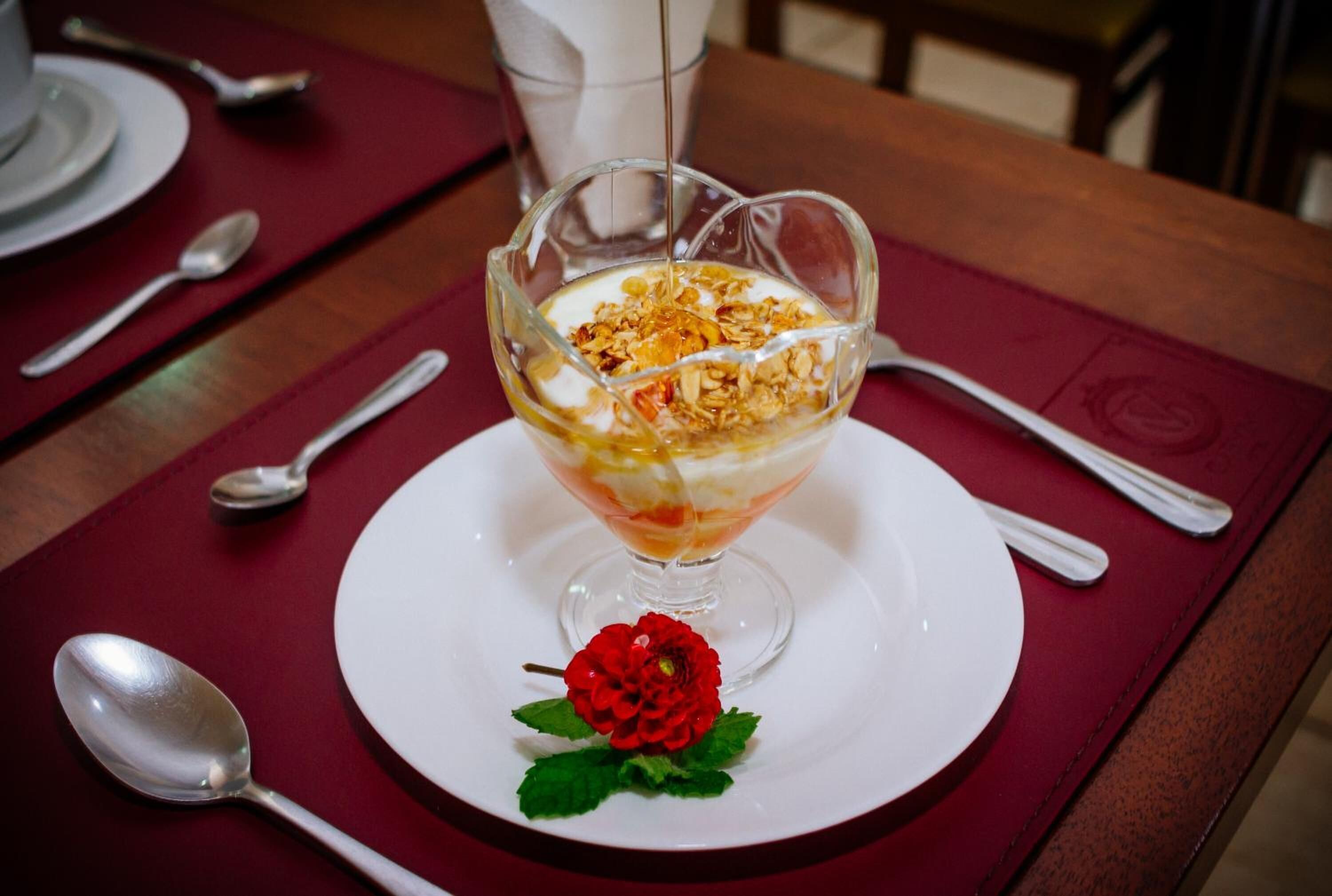 Un dessert servi dans un bol en verre, posé sur une assiette blanche et agrémenté d’une rose rouge, le tout sur une nappe rouge.