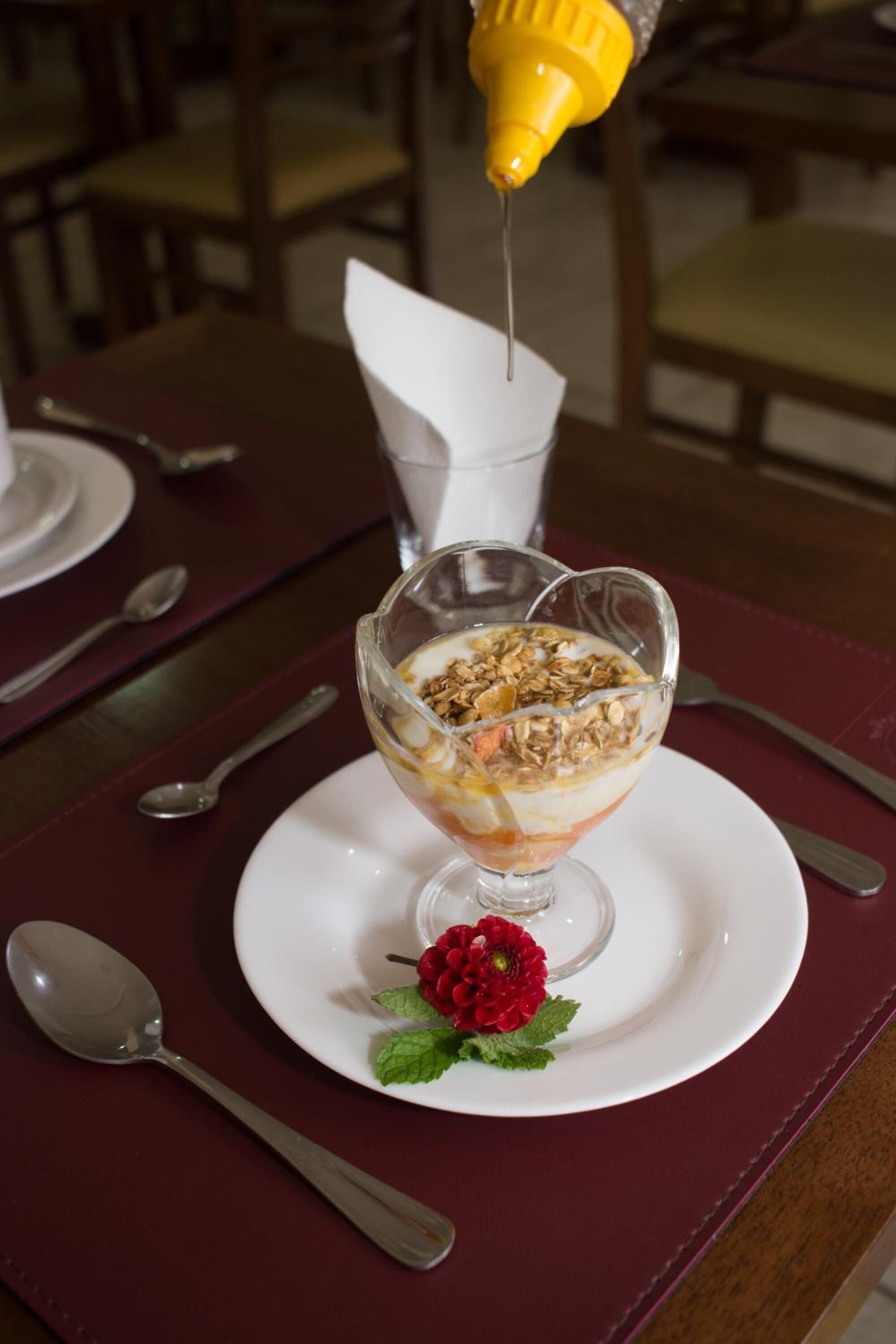 Dessert in einer Glaskugel auf einem weißen Teller, garniert mit einer roten Blüte, mit Löffel und Servietten auf einem Tisch.
