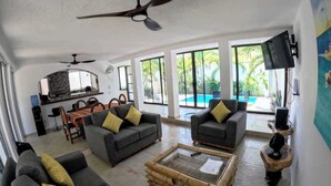 Ein geräumiger Wohnbereich mit zwei Sofas, einem Couchtisch und einem Deckenventilator. Im Hintergrund ist ein Essbereich zu sehen und ein großes Fenster mit Blick auf einen Pool.