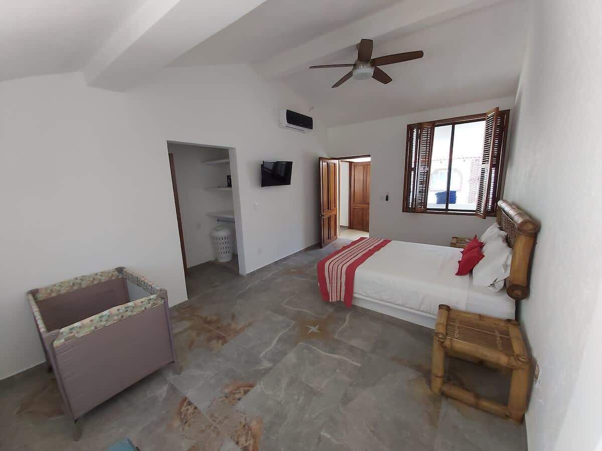 Une pièce comprenant un lit, une chaise en bois, un ventilateur de plafond et une fenêtre avec des volets.
