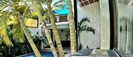 Un área de piscina moderna con suelo de baldosas, palmeras y una puerta de cristal que da a un porche cubierto.