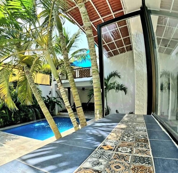 A modern pool area with a tiled floor, palm trees, and a glass door leading to a covered patio.