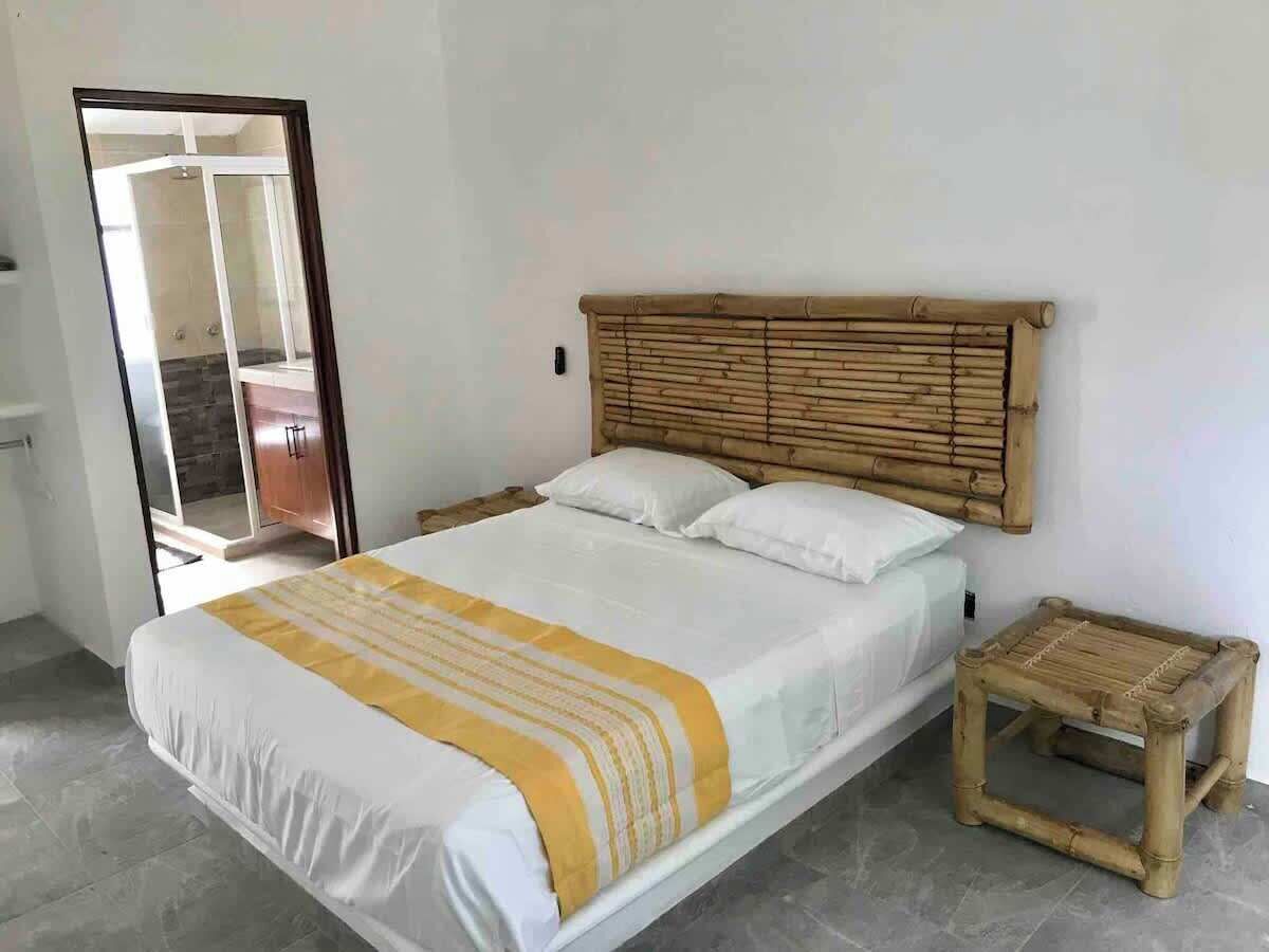 Un dormitorio con cama, cabecero de madera, mesita de noche y baño visible a través de una puerta de cristal.