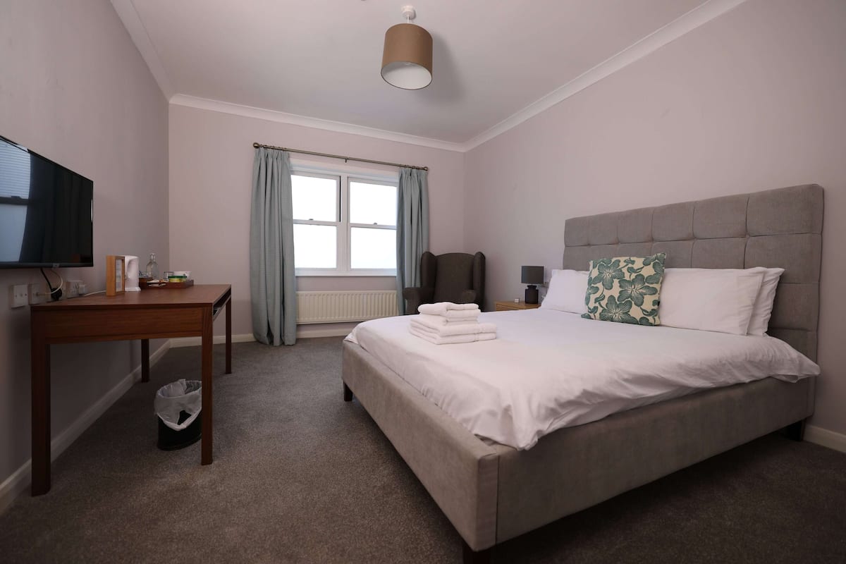A bedroom with a bed, a desk, a television, a window with curtains, and a ceiling light.
