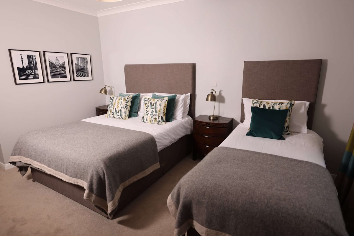A hotel room with two beds, each with a grey bedspread and matching headboards. There are three framed pictures on the wall, a bedside table with a lamp, and a small drawer unit.