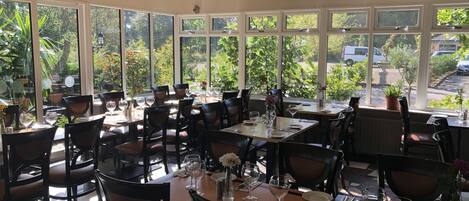A restaurant with multiple tables and chairs, large windows offering a view of greenery and parked cars outside.