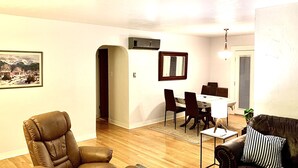 A living room with a leather armchair, a sofa with a striped pillow, a dining table with chairs, and a framed picture on the wall.
