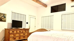 A bedroom with a wooden bed, a TV on a dresser, and white blinds on the windows.
