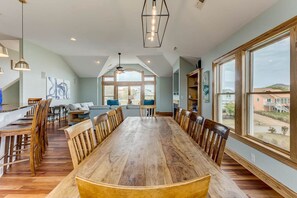 Un comedor amplio con una larga mesa de madera rodeada de sillas, ventanas grandes que ofrecen vistas al exterior, y una cocina moderna al fondo.
