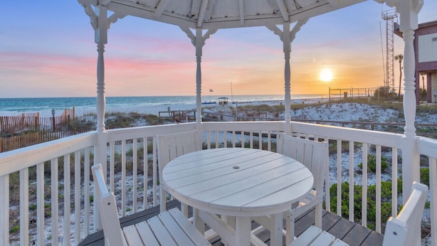 Um gazebo com uma mesa redonda e bancos, com vista para uma praia ao pôr do sol.