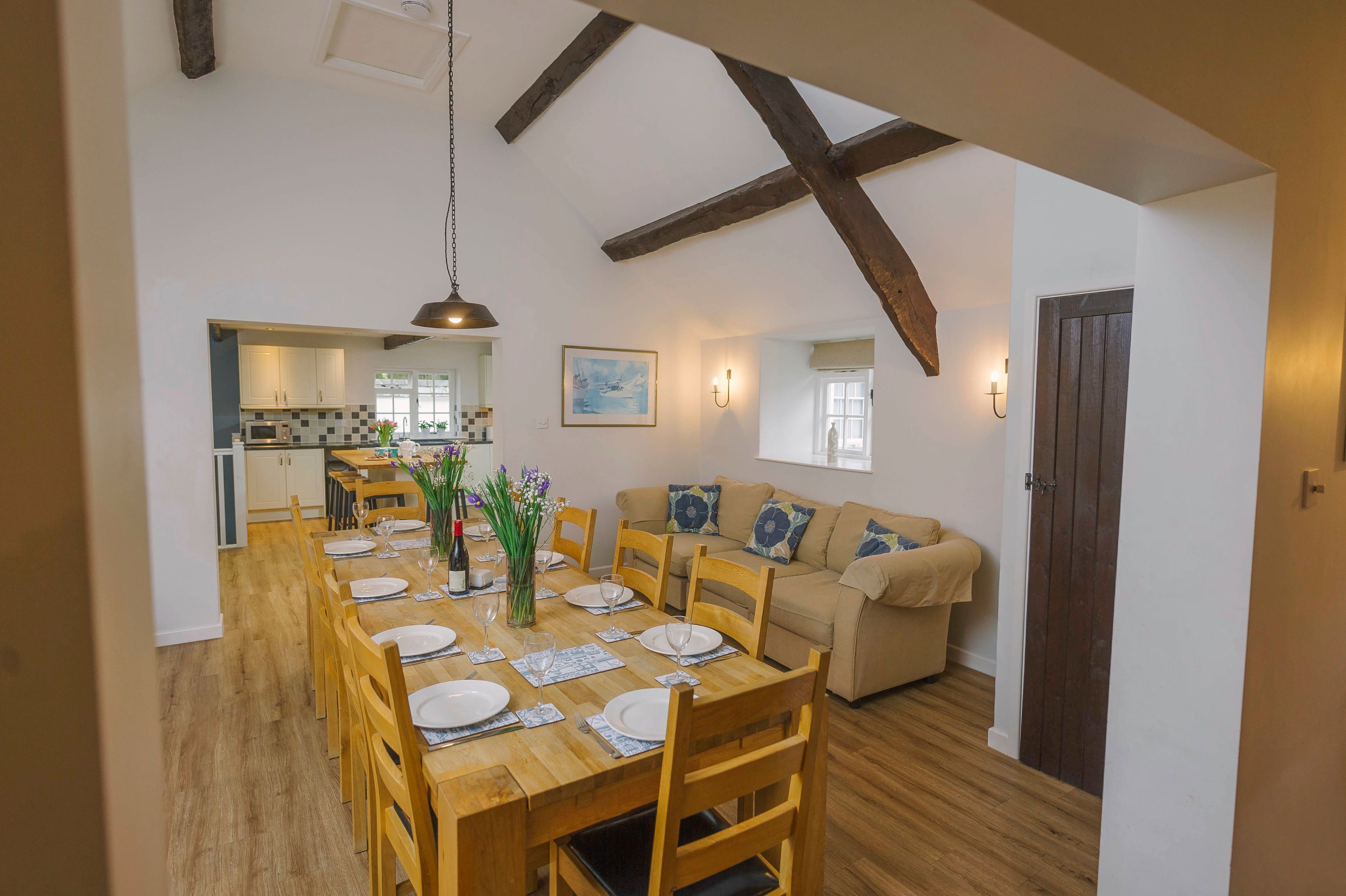Zona pranzo con tavolo in legno apparecchiato per sei persone, un divano e una cucina sullo sfondo.