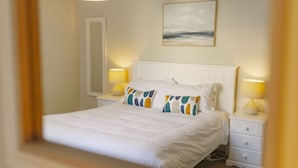 A neatly made bed with white bedding and colorful pillows, two bedside lamps, a nightstand, a mirror, and a painting of a beach scene on the wall. - Corffe Cottages, Perfect Devon retreat, sleeps 4, heated indoor pool (Barnstaple)