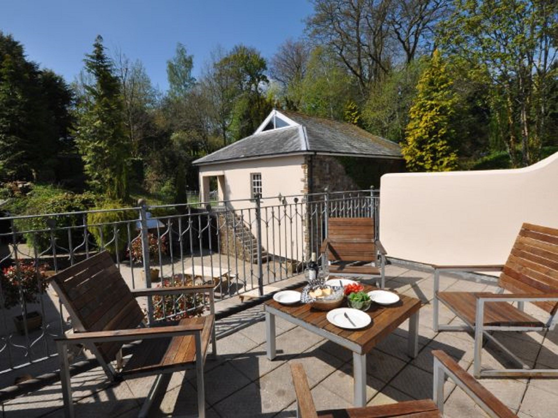 Une terrasse avec des meubles d’extérieur, une petite table dressée avec des assiettes et un saladier rempli de fruits, et un bâtiment en pierre en arrière-plan.
