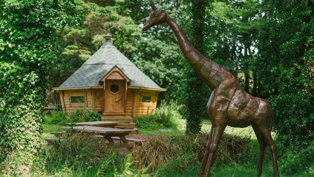 A wooden cabin with a thatched roof surrounded by lush greenery and a giraffe statue. - Corffe Cottages, Perfect Devon retreat, sleeps 4, heated indoor pool (Barnstaple)