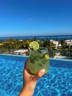 Eine Hand hält ein Glas mit einem Limettenschnitz und Minzblättern, mit Blick auf einen Pool mit klarem, blauem Wasser und im Hintergrund Meer und Palmen.