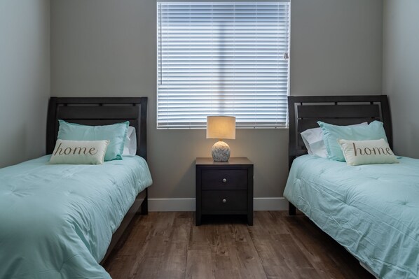 A bedroom with two beds, each with a pillow labeled "home," a nightstand with a lamp, and a window with blinds. - Grill Deck + Game Lounge | Mins to Downtown Paso | Westside Riley House (Paso Robles)