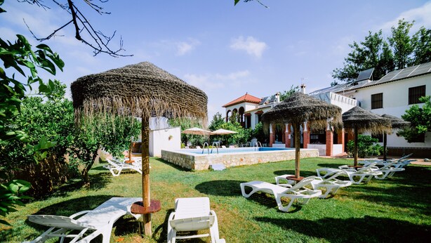 A pool area with lounge chairs and thatched umbrellas, surrounded by greenery and a building with a red roof. - San Antonio Rural House. Villa 3 (3 bedrooms) (Cabra (Córdoba))