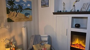 Ein gemütliches Wohnzimmer mit Kamin, einem Strandbild und einem bequemen Sessel.