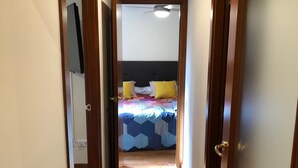 Un couloir étroit mène à une chambre à coucher équipée d’un lit, d’un ventilateur de plafond et d’un téléviseur fixé au mur.