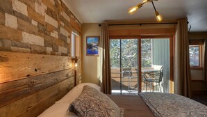 A bedroom with a wooden headboard, a bed with patterned bedding, a ceiling light fixture, and a balcony with a table and chairs.