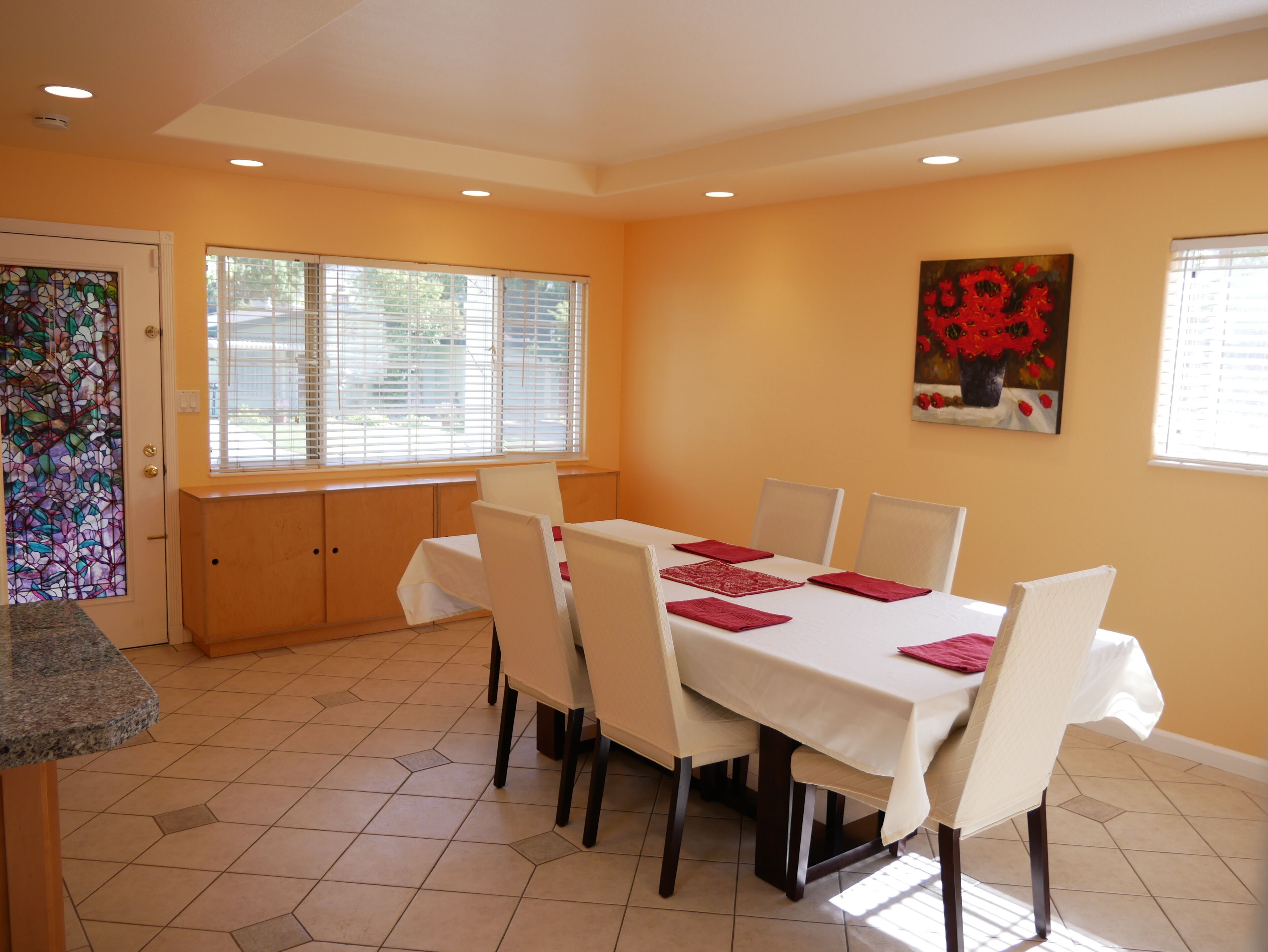 A dining area with a table set for six, white chairs, and a painting of red flowers on the wall.