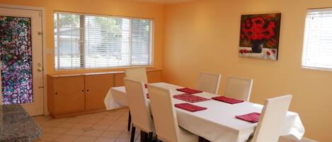 A dining area with a table set for six, white chairs, and a painting of red flowers on the wall.