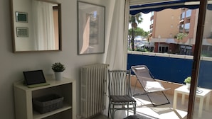 Un balcon avec une chaise, une table, un radiateur, une étagère avec des paniers et une plante en pot.