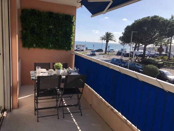 A balcony with a table set for dining, a potted plant, and a view of the sea and parked cars. - Beautiful 2 bedroom apartment by the sea (4 pers) (Fréjus)