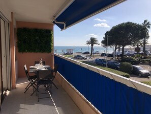 Un balcón con mesa dispuesta para comer, vista al mar y coches aparcados.