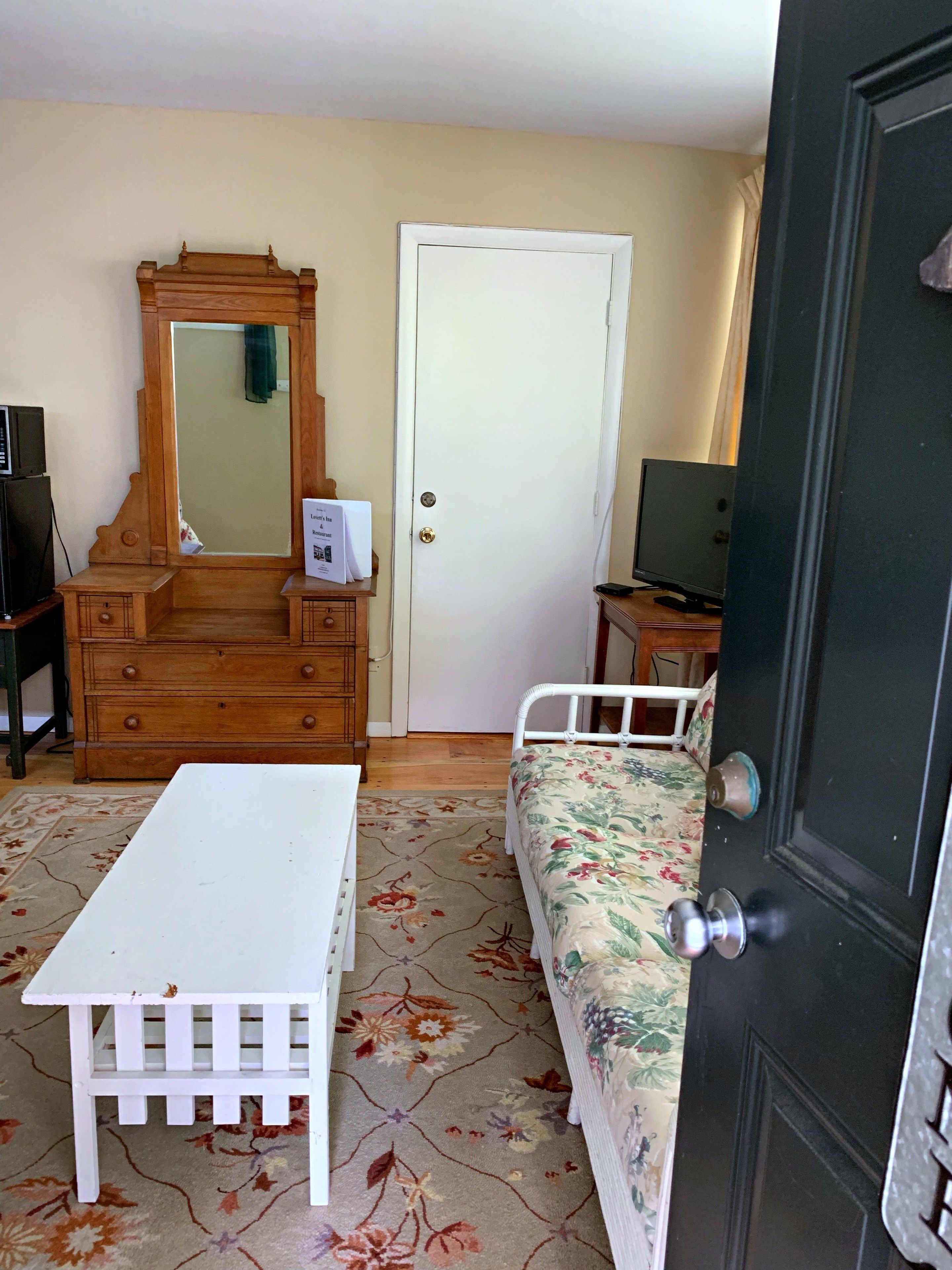 A bedroom with a wooden dresser, a white bedside table, a floral-patterned bed, a mirror, a door, and a television.