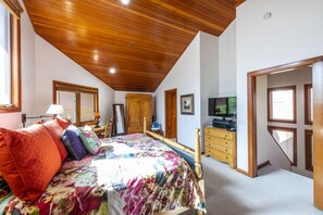 Un dormitorio con techo de madera, una cama con ropa de cama colorida, un aparador, un televisor y un espejo.