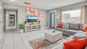 A modern living room with a grey sofa, a coffee table, and a TV. There is a door leading to a laundry room, and a window with curtains. - 🌞6 KING beds, 14 GUESTS, 5 BEDROOMS, 2 BATHROOMS, REFRIGERATION, NEAR AIRPORT (El Paso)