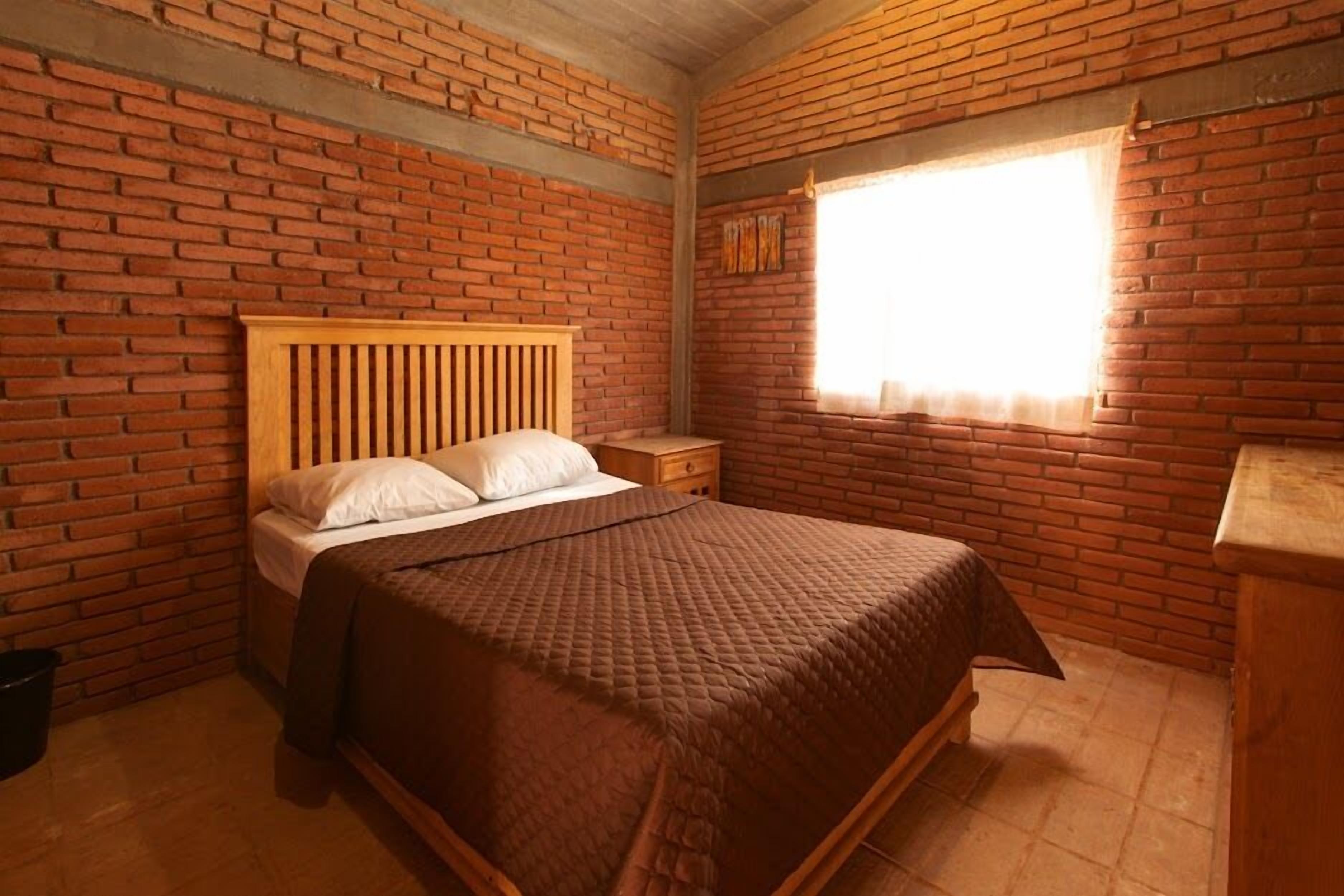 A room with a brick wall, a wooden bed, a nightstand, and a window with curtains.