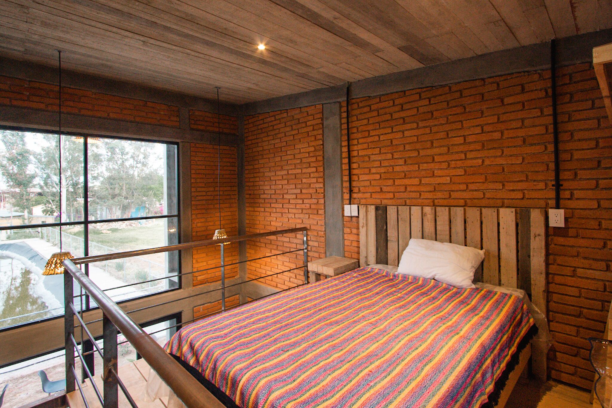 A room with a brick wall, a bed with a colorful blanket, a wooden headboard, and a window with a view of trees.