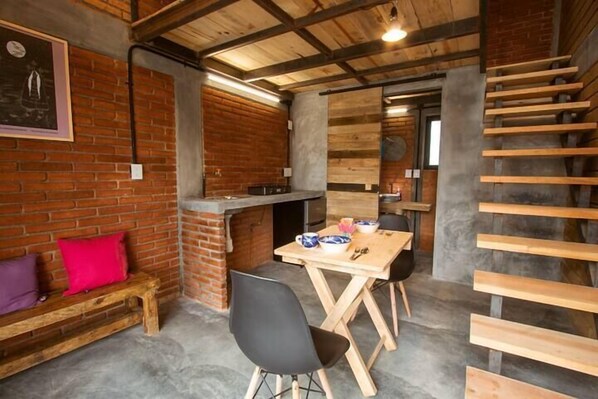 Un interior rústico con paredes de ladrillo visto, un banco de madera, una mesa de comedor con sillas y una escalera de madera.
