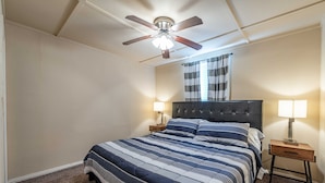 Ein Schlafzimmer mit einem Bett, zwei Nachttischen, einem Deckenventilator, einem Fenster mit Vorhängen und einem an der Wand befestigten Spiegel.