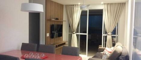 A dining area with a red table, grey chairs, and a ceiling fan. There is a sofa with cushions and a window with curtains.