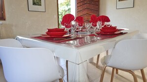 Una mesa de comedor dispuesta con servilletas rojas, platos y vasos, rodeada de sillas blancas en una habitación con cuadros en las paredes.