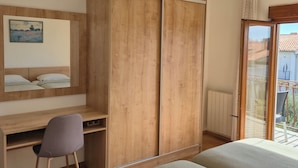 A bedroom with a wooden wardrobe, a desk with a chair, a bed, and a view of the outside through a glass door.